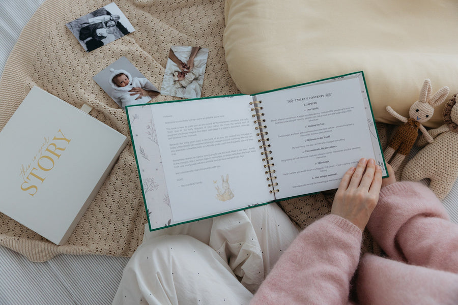 The CheriBOO Baby Memory Book in Evergreen, open at its table of contents on a knitted blanket, with printed photos and the keepsake box beside it