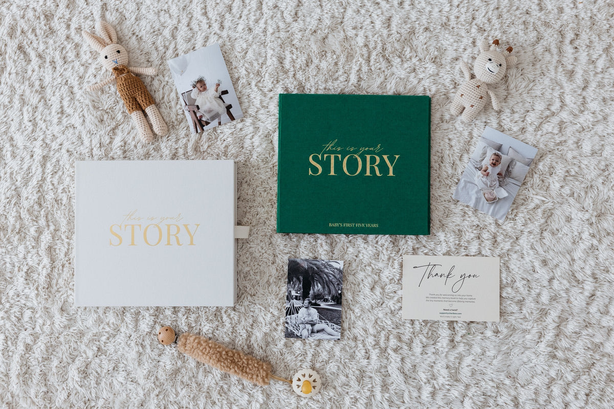 Everything in the box: the CheriBOO Baby Memory Book in Evergreen, its keepsake box, thank-you card and printed photos laid out on a rug