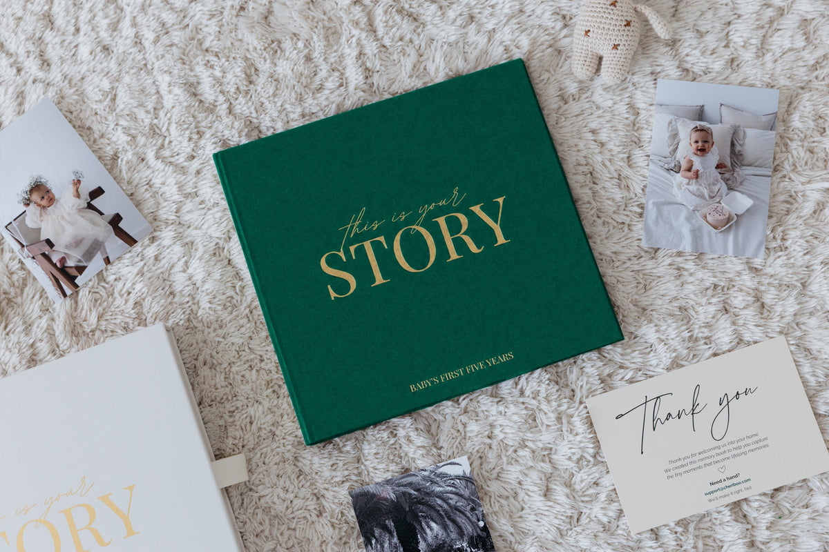 Close-up of the CheriBOO Baby Memory Book in Evergreen with its gold foil title, next to printed photos and the keepsake box