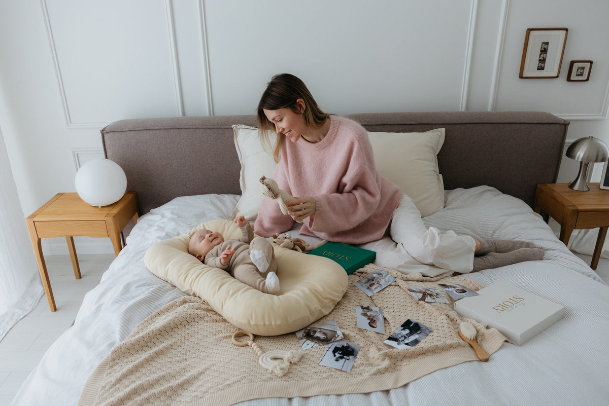 A mother plays with her baby on the bed; the CheriBOO Baby Memory Book in Evergreen and its keepsake box lie beside printed photos