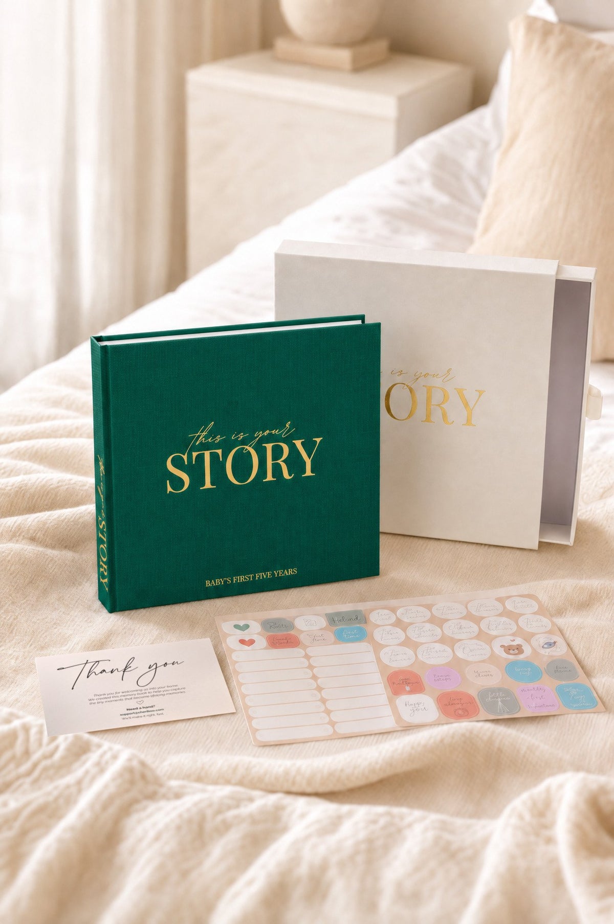 The CheriBOO Baby Memory Book in Evergreen with its keepsake box, thank-you card and sticker sheets on a bed