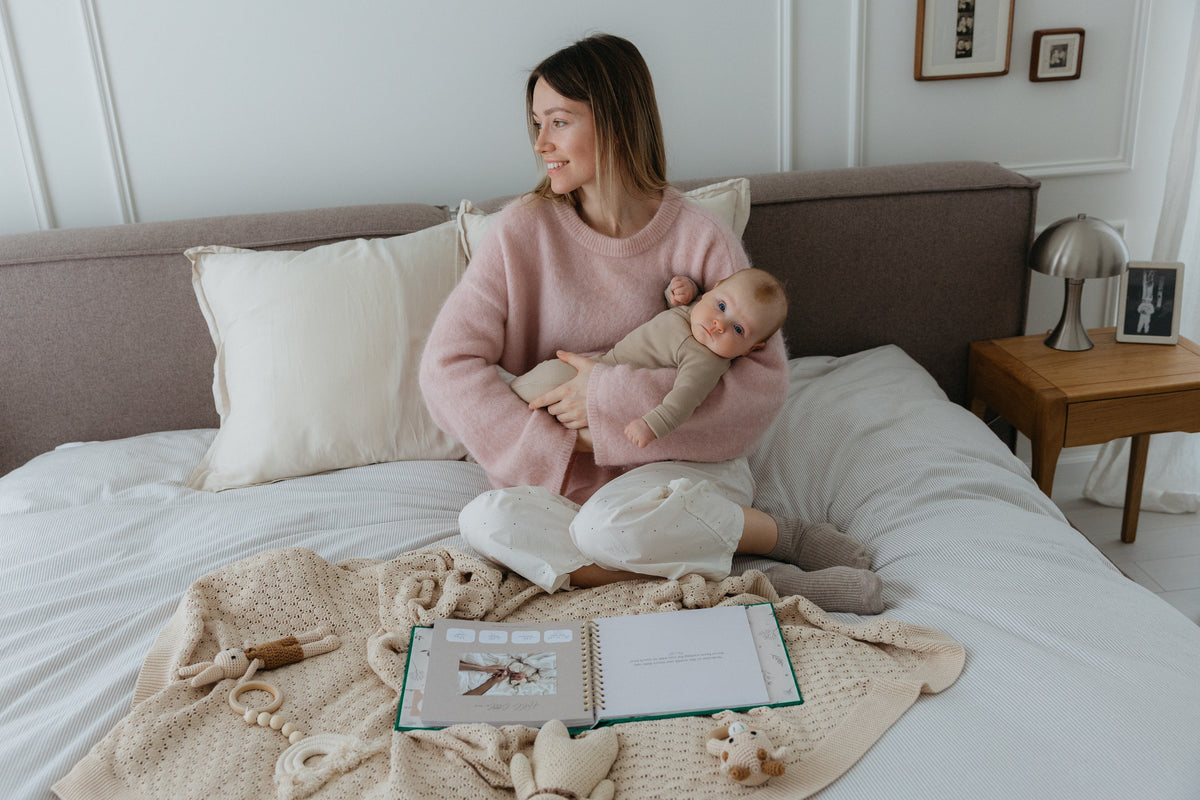 A mother holds her baby on the bed with the CheriBOO Baby Memory Book open in front of them