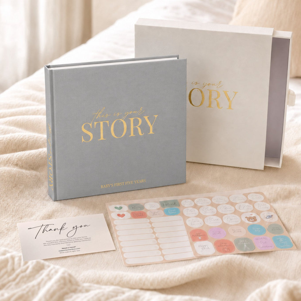 The CheriBOO Baby Memory Book First Edition in Light Gray with its keepsake box and sticker sheets on a bed