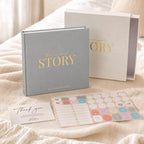 The CheriBOO Baby Memory Book First Edition in Light Gray with its keepsake box and sticker sheets on a bed