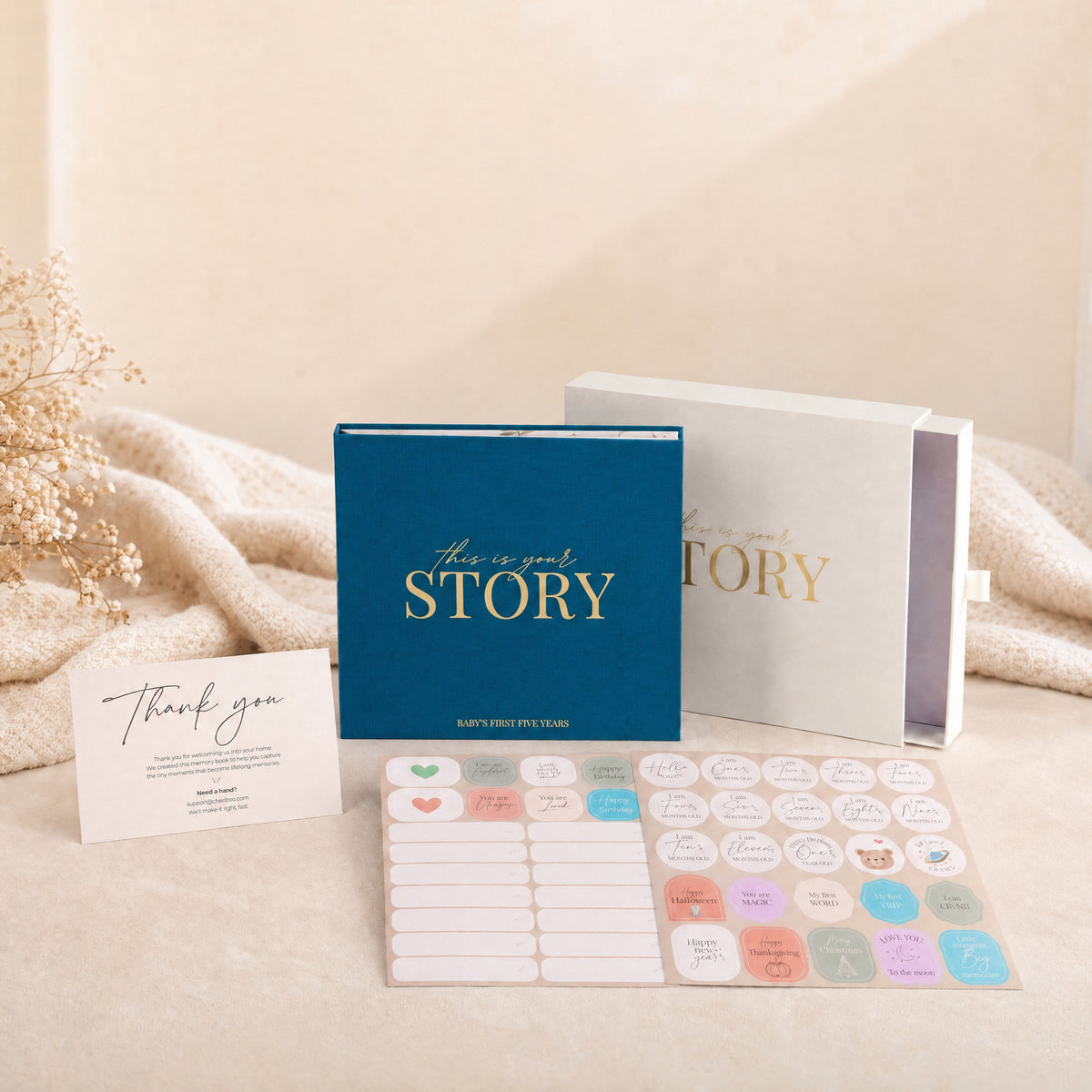 The CheriBOO Baby Memory Book in Deep Sea Blue with its keepsake box, thank-you card and two sticker sheets on a linen throw