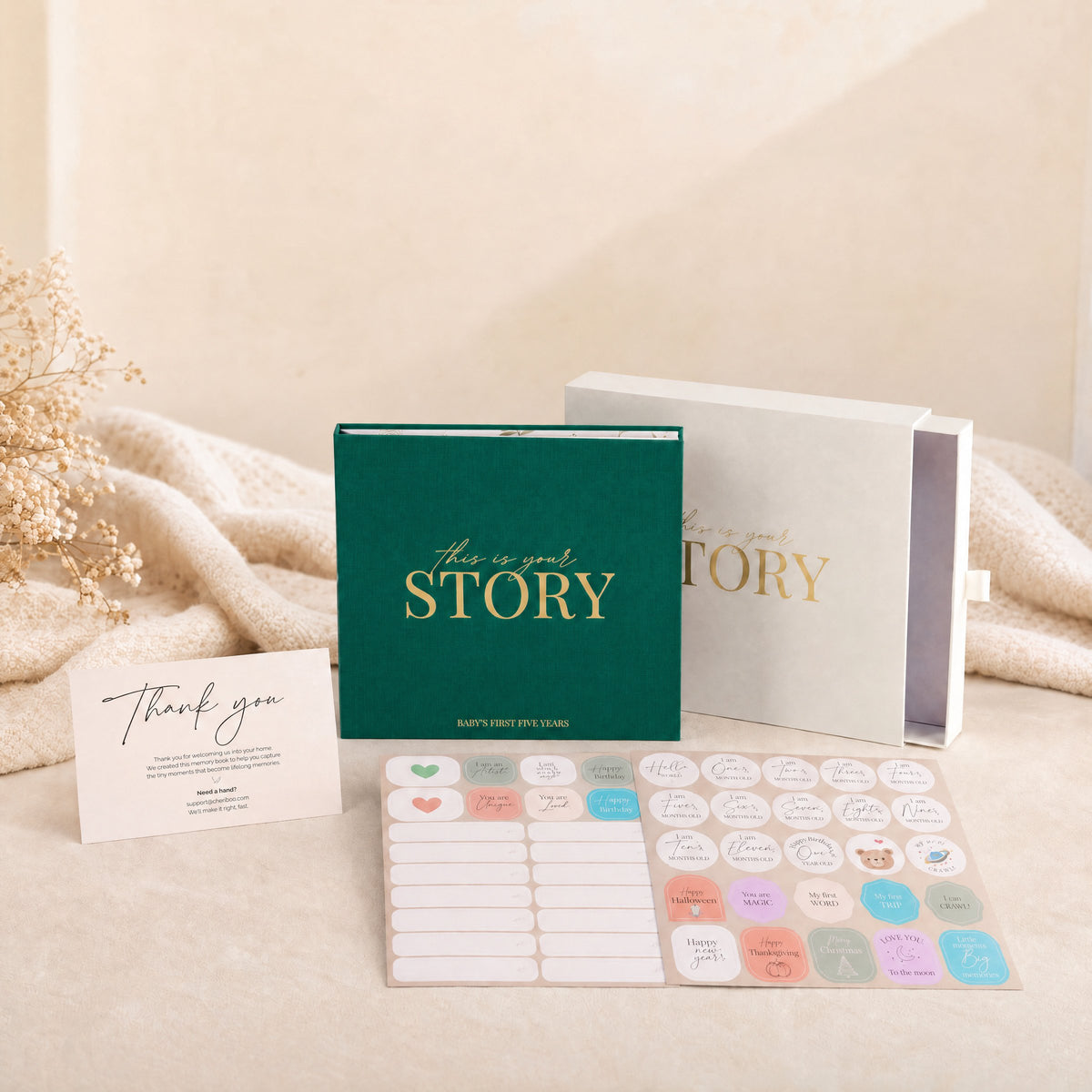 The CheriBOO Baby Memory Book in Evergreen with its keepsake box, thank-you card and two sticker sheets on a linen throw
