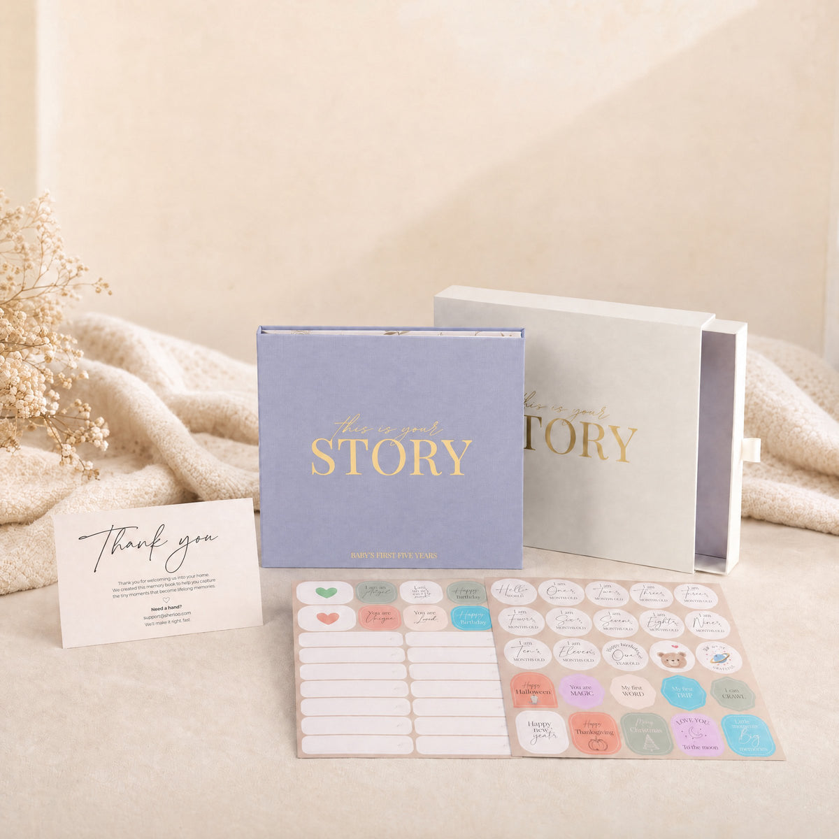 The CheriBOO Baby Memory Book in Lavender with its keepsake box, thank-you card and two sticker sheets on a linen throw