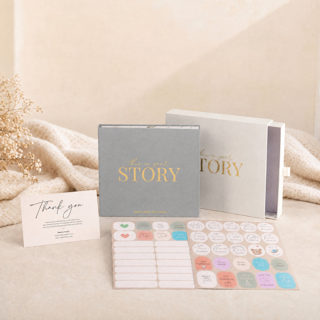 The CheriBOO Baby Memory Book First Edition in Light Gray with its keepsake box, thank-you card and two sticker sheets on a linen throw